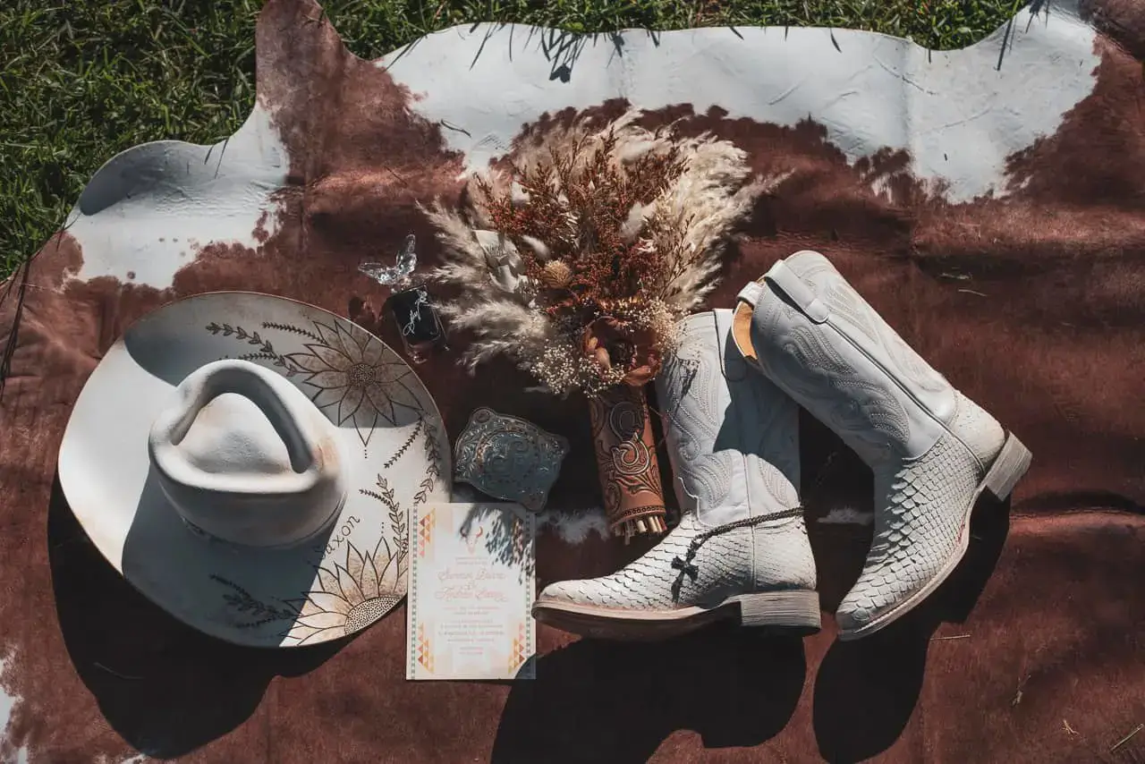 Flat lay of white cowboy boots, a custom floral cowboy hat, dried floral bouquet, and wedding invitation on a brown and white cowhide rug outdoors.
